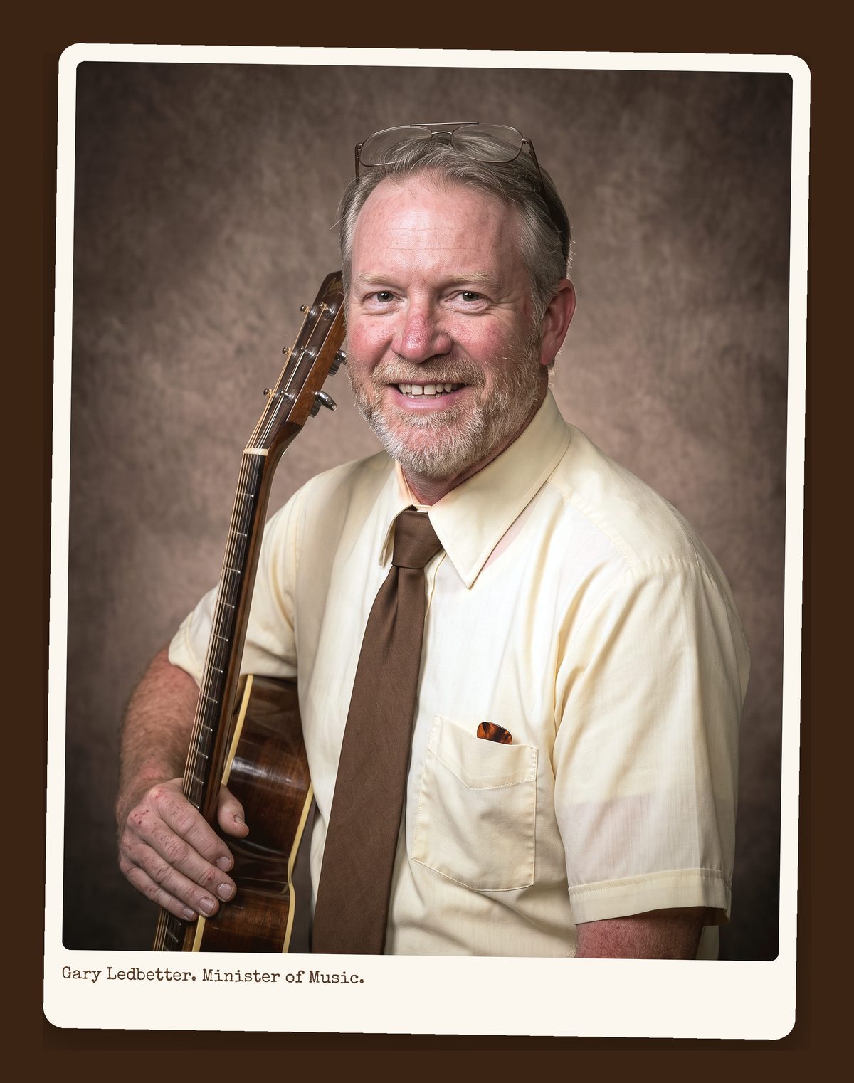 Gary Ledbetter, graying beard, short-sleeve shirt and brown tie, holding an acoustic guitar, directory portrait.