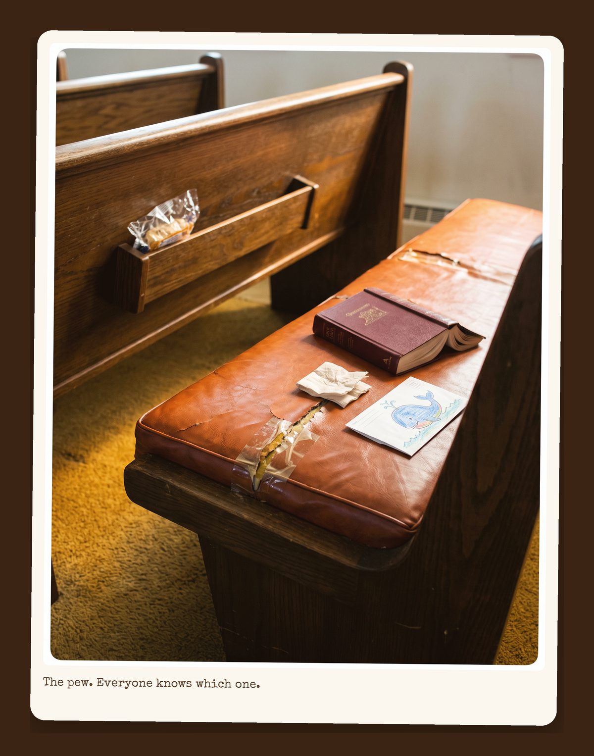 One wooden pew with an orange cushion, a burgundy hymnal, a peppermint, and a strip of tape at a seam in the cushion.